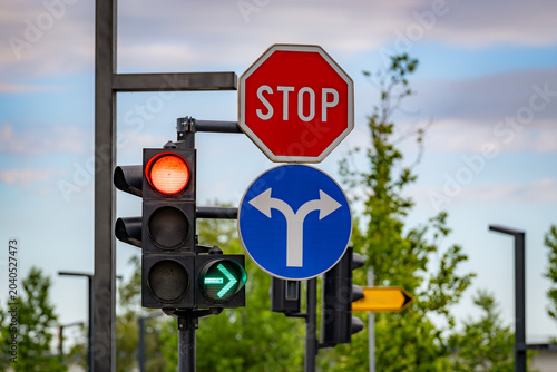 Stop sign, blue direction sign, and traffic lights at a modern city intersection with trees and sky in the background.