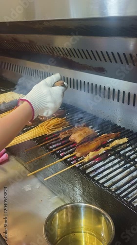 A chef grills skewers of marinated meat, mushrooms, and other ingredients over charcoal on a commercial barbecue grill, with smoke rising from the fire at vertical composition