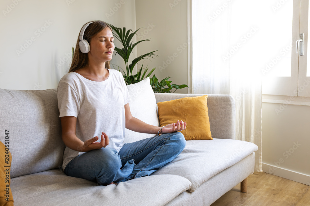 Fototapeta premium Young woman meditating on the couch listening to guided meditation using wireless headphones. Copy space.