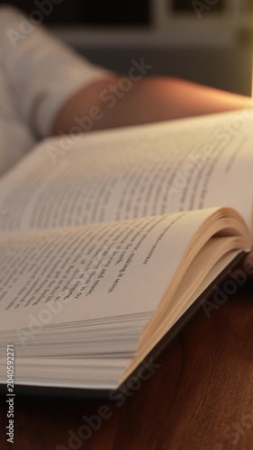 A woman's hand turns pages of an open book resting on a dark wooden table, bathed in warm golden lamp light, creating a cozy intimate reading atmosphere with soft bokeh in the background.