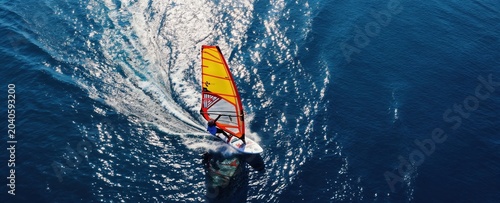 Windsurfer rides fast across deep blue ocean water creating white splash waves. Yellow sail catches wind power. Drone view from high above shows surfer on surfboard moving at speed.