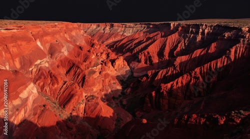 Vast red rock canyon landscape with dramatic shadows. Steep, eroded cliffs display stratified rock layers. Barren desert terrain borders dark sky.