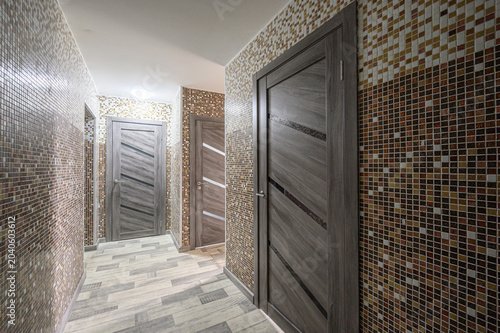 A narrow hallway features walls covered in multicolored glass mosaic tiles, grey wood-textured doors, and a light grey wood-patterned floor, creating a modern and textured interior