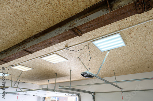 OSB-paneled garage ceiling with LED square lights; rusty steel beam runs along the top, automatic garage door opener and metal tracks visible below
