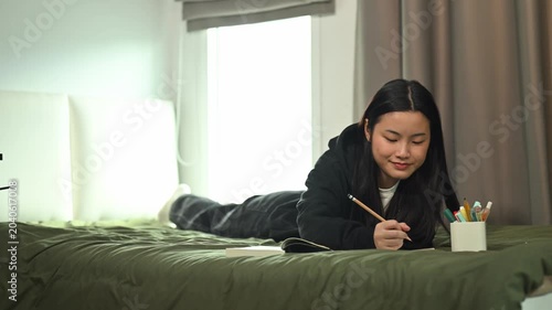 Young Asian Female Student Writing in Tablet on Bed in Cozy Dorm Room by Window