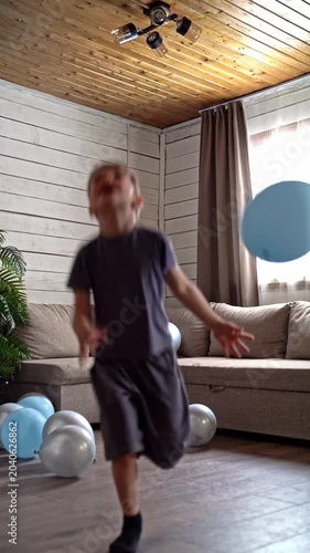 Boy playing alone with floating balloon, energetic child chases and juggles blue balloon among scattered silver ones, sunlight through curtains and wooden ceiling highlight freedom and motion