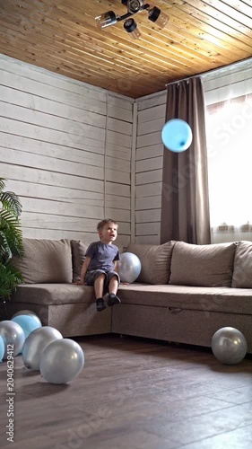 Young boy tossing blue balloon across cozy living room, jumping from sofa, reaching for floating balloon with giggle and energy, scattered silver balloons on hardwood floor, warm sunlight