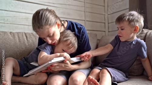 White mother comforting son between pages on couch while younger white child flips album, soft gestures and close embrace create symbolic reassurance, gentle mood of protection and shared family