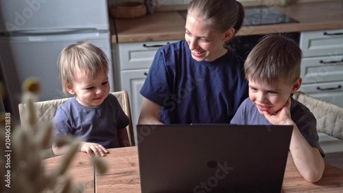 White mother and sons smiling at laptop together in kitchen, shared laughter, tech bridging distance, warm emotional connection, candid moment, playful reactions, sunlight warmth, gentle eye contact,