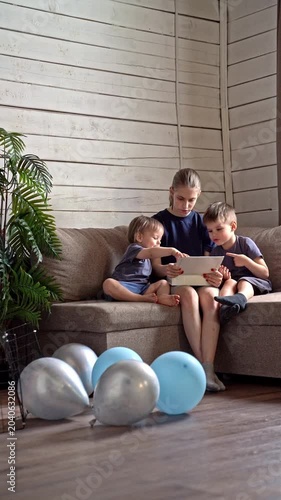 Siblings learning together. Young sister guides brothers through educational activity on sofa. Playful scene of sister instructing brothers on tablet amid cheerful home decor and decorations