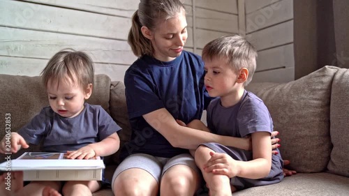 Caucasian mother on couch with tablet guiding two young sons through picture exploration, wooden wall backdrop, casual clothing, close embrace, patient instruction, focused toddler tapping screen,