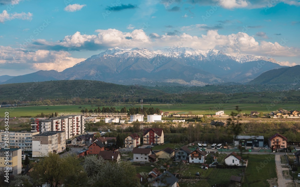 Fototapeta premium Breathtaking scenery unfolds as snow-capped mountains rise above a charming village during dusk. Homes dot the landscape while lush fields stretch towards the horizon, creating tranquility