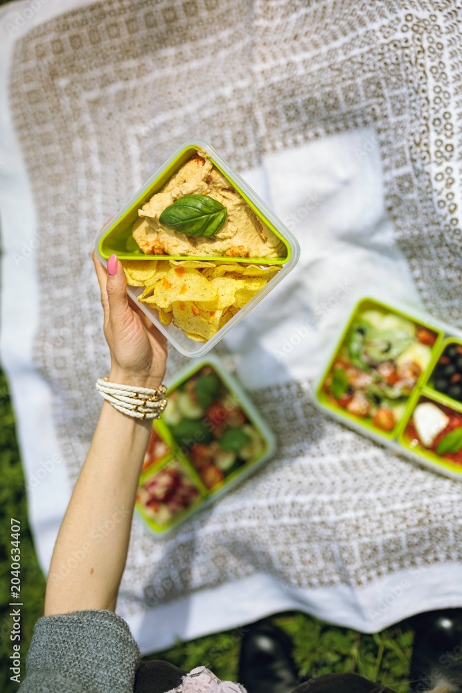 Fototapeta premium A hand holds a green container of chips and dip, surrounded by vibrant salads and snacks laid out on a white blanket in a sunny park. A perfect day for outdoor dining