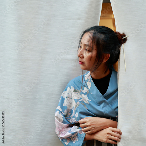 Japanese woman in blue floral kimono peeking through white noren curtain, Kyoto, Japan