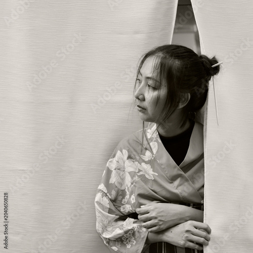 Japanese woman in blue floral kimono peeking through white noren curtain, in sepia black and white, Kyoto, Japan