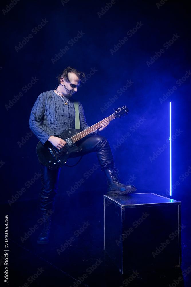 Obraz premium Adult man playing black electric guitar with foot on box in dark smoky stage with blue neon light