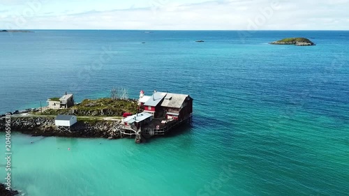A small village sits by the water. Houses and trailers line the coast. Green hills rise in the background under a clear sky. The calm water reflects the scenery.