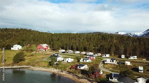 A small village sits by the water. Houses and trailers line the coast. Green hills rise in the background under a clear sky. The calm water reflects the scenery.