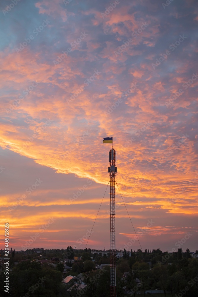 Obraz premium Telecommunications tower against the backdrop of a cityscape at sunset, Ukraine. Ukrainian flag on a telecommunications tower at sunset.