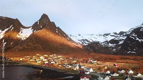 A small town is visible along the coast of Norway. Mountains rise in the background. Sunlight casts a warm glow on buildings. The sky remains mostly clear as the day ends.