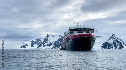A large cruise ship floats in the cold waters of Antarctica surrounded by snow-covered mountains under gray clouds. Small icebergs are visible in the distance.