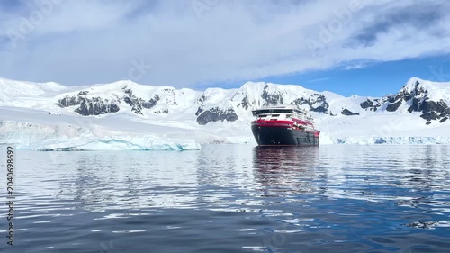A large cruise ship floats in the cold waters of Antarctica surrounded by snow-covered mountains under gray clouds. Small icebergs are visible in the distance.