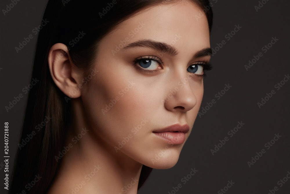 Fototapeta premium Close-up portrait of young woman with dark hair and blue eyes. Face with smooth skin and natural makeup. Head turned sideways, looking at camera. Studio shot, dark background, copy space.