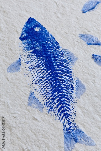 Abstract gyotaku style fish print in blue ink on textured white paper, showcasing intricate details of scales and fins with additional smaller fish impressions surrounding it