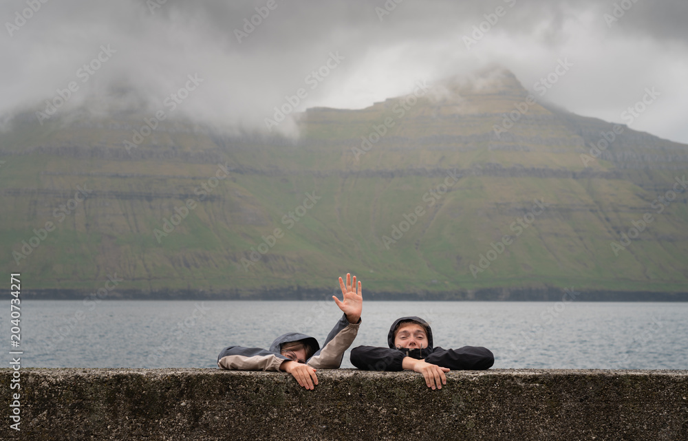 Fototapeta premium Humorous concept of a young couple in raincoats pretending to sink and reaching for help against misty mountains in the Faroe Islands