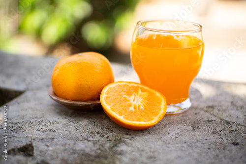 Fresh orange juice in a glass with halved and whole oranges on a stone ledge, bright outdoor bokeh background