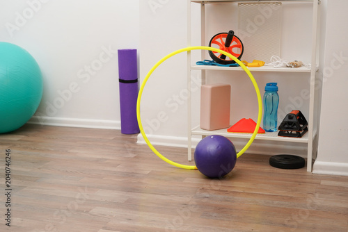 A variety of colorful fitness equipment arranged for a home workout. Includes a stability ball, yoga mat, hoop, and weights on a wooden floor. Bright, clean setting for a healthy lifestyle.