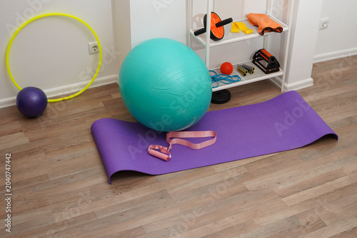 A collection of colorful exercise equipment for a home workout or physical therapy. A purple yoga mat, turquoise stability ball, and resistance bands are on a wooden floor in a bright, modern room.