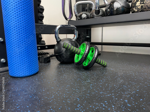 A variety of functional training equipment ready for a workout. A green ab wheel, black kettlebell, and blue foam roller sit on a black rubber floor in a modern gym. Bright and clean lighting.