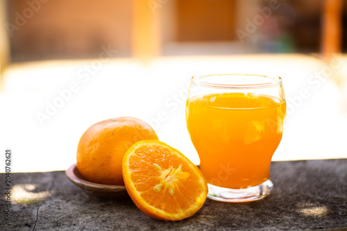 Fresh orange juice in a glass with halved and whole oranges on a stone ledge, bright outdoor bokeh background