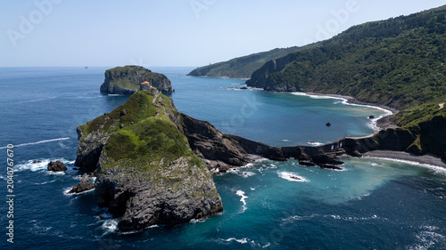 Aerial drone view of Gaztelugatxe, a small rocky island connected to the mainland by a historic stone bridge and staircase. Blue waters of the Bay of Biscay. Impressive cliffs, rugged landscapes, and 