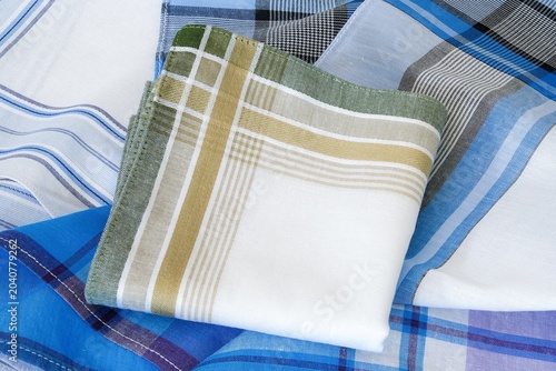 A neatly folded white cotton handkerchief featuring olive green and mustard yellow striped borders is elegantly displayed on unfolded men's cotton handkerchiefs.