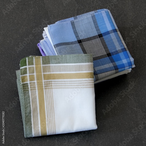 A white handkerchief with green and gold stripes sits folded on top of an assortment of blue and plaid cotton men's handkerchiefs.