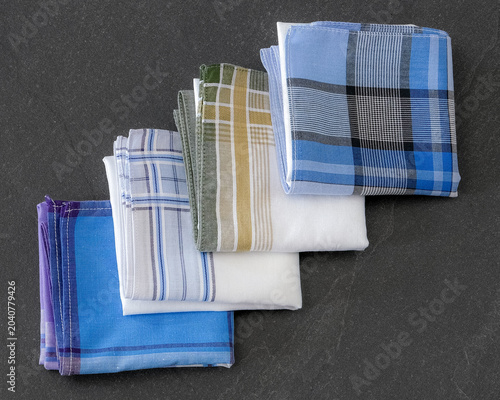 Four diverse folded cotton handkerchiefs in shades of blue, purple, white, and green on a textured dark background.