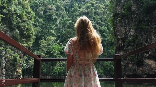 Woman in floral dress walking to wooden railing to look at tropical jungle lake and limestone cliffs during daylight. Back view of female traveler enjoying nature scenery in Southeast Asia.
