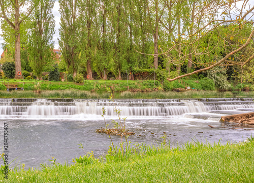 A weir at a riverside park with a line of trees in the background.