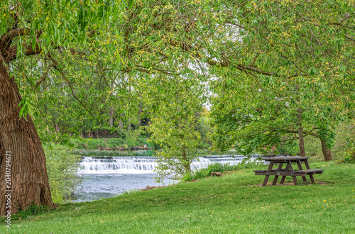 A weir at a riverside park with a line of trees in the background.