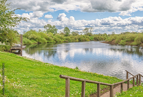 A river meanders through a rural setting with a tree-lined riverbank. A church spire is on the skyline and a sky with cloud is above.