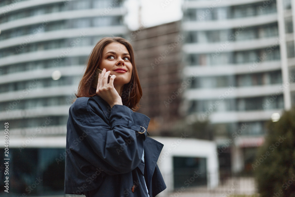 Fototapeta premium A thoughtful woman in a coat stands outdoors in an urban cityscape, talking on a phone, with modern buildings behind, capturing casual communication in daily life.