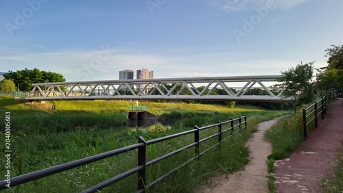 Canvastavla pedestrian truss bridge over a riverside trail in an urban green space