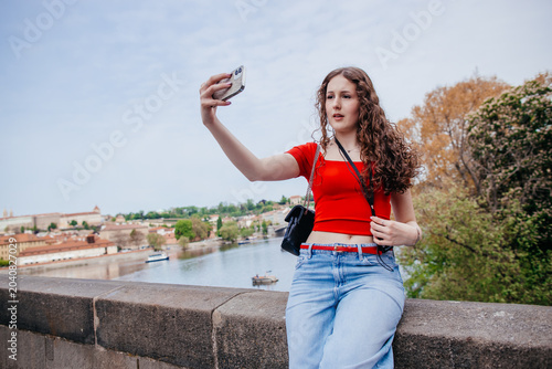 Young woman taking selfie with smartphone while sitting on bridge overlooking river city