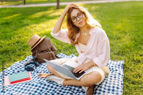 A woman sits on a blanket in a park, working on a laptop. A female freelancer enjoys the sunset outdoors with a laptop. Conceptual image of technology, leisure, and education.