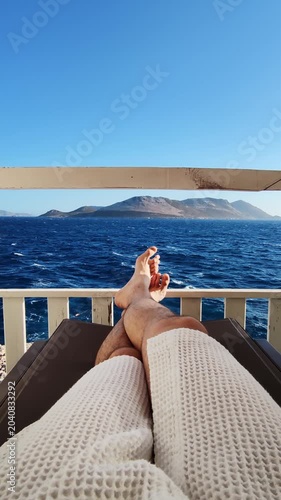 Low section of man legs lying on sunbed situated on dock with serene seascape in slow motion. male caucasian person in bathrobe enjoying the view of ocean waves. spending tranquil time on vacations