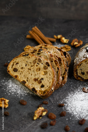 Sliced raisin walnut bread on a dark slate with cinnamon sticks, walnuts and powdered sugar, rustic homemade bakery on a dark background