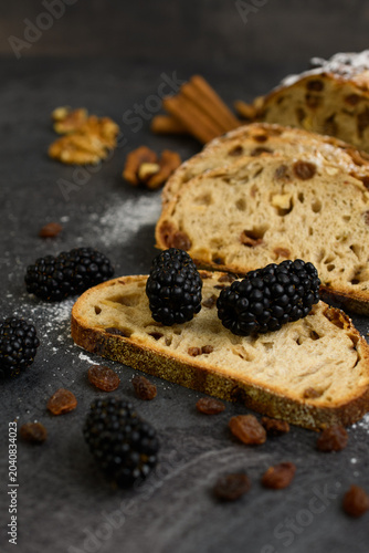 Slices of raisin walnut bread topped with fresh blackberries, cinnamon and walnuts dusted with powdered sugar on a dark rustic background