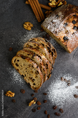 Sliced raisin walnut bread on a dark slate with cinnamon sticks, walnuts and powdered sugar, rustic homemade bakery on a dark background